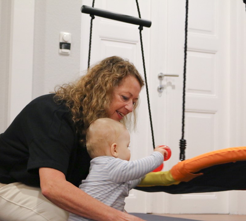 Doris Godt mit einem Säuglingspatienten vor einer Schaukel in ihrer Physiotherapiepraxis
