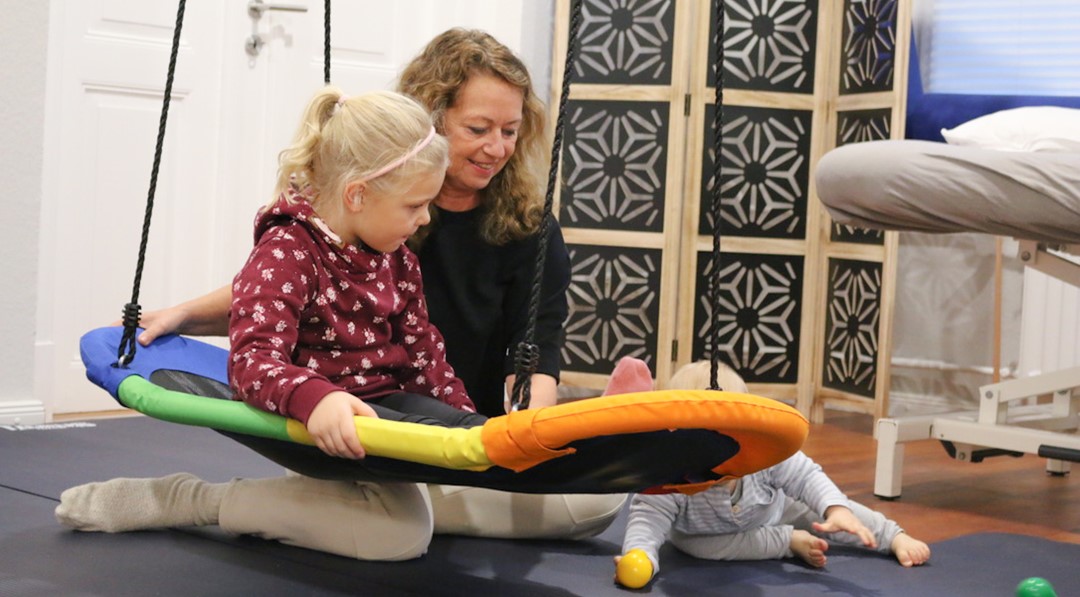 Doris Godt mit zwei Kindern in ihrer Physiotherapiepraxis, eins sitzt auf einer Schaukel, eines krabbelt dahinter